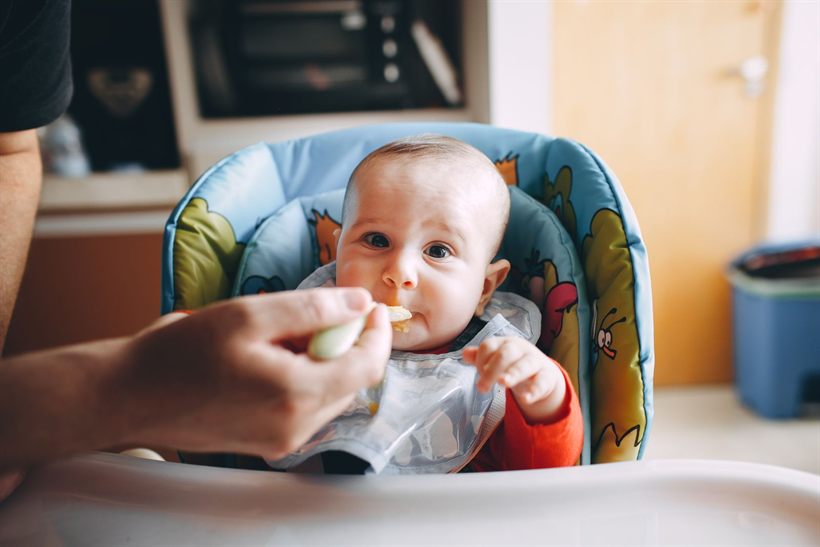 Parent spoon-feeding a six-month-old baby in a high chair
