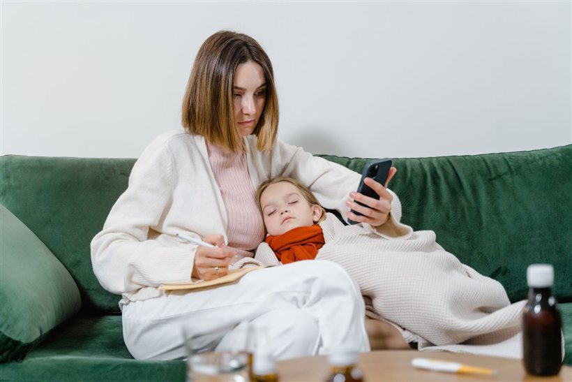 Mother checking symptoms on her phone while her sick child rests on the couch